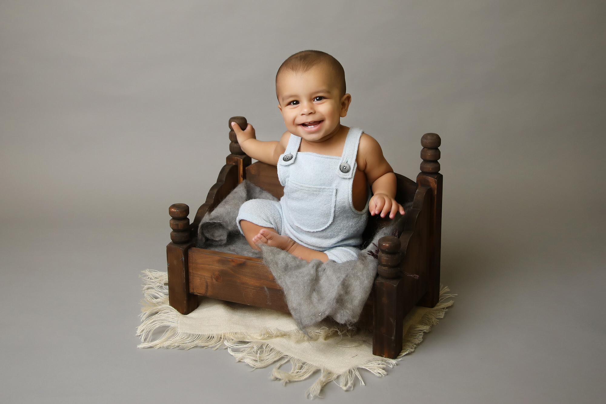 baby boy sitting in a wooden bed, wearing blue dungarees, by Nursery Photography in Durham